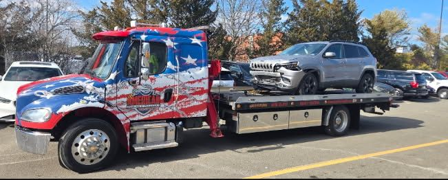 Tow truck hauling a damaged silver SUV, truck features American flag graphics.