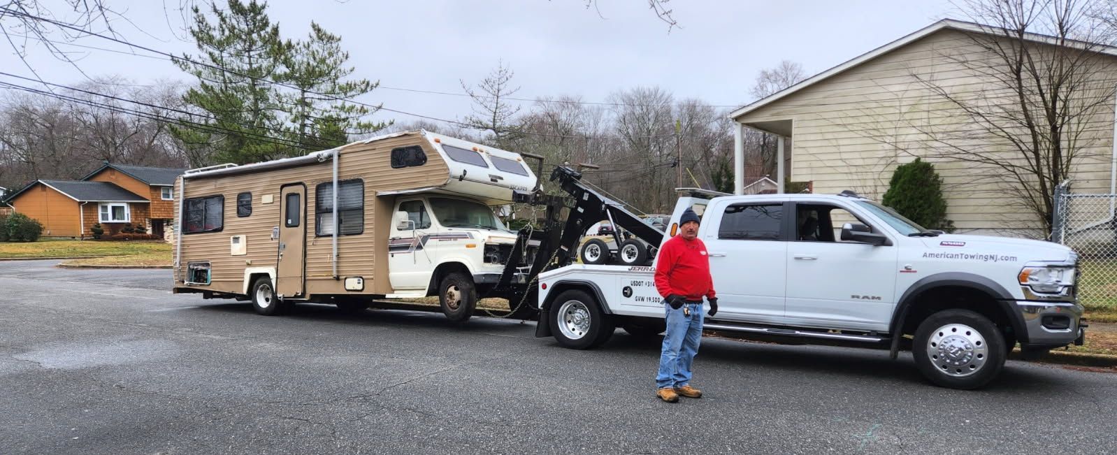 A white tow truck is towing a brown and white RV on a residential street. A person in a red jacket stands beside the truck.