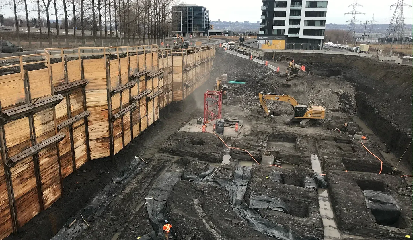 Un chantier de fouilles avec des murs de soutènement en bois, une excavatrice jaune et des coffrages de fondation en béton sur un sol boueux.