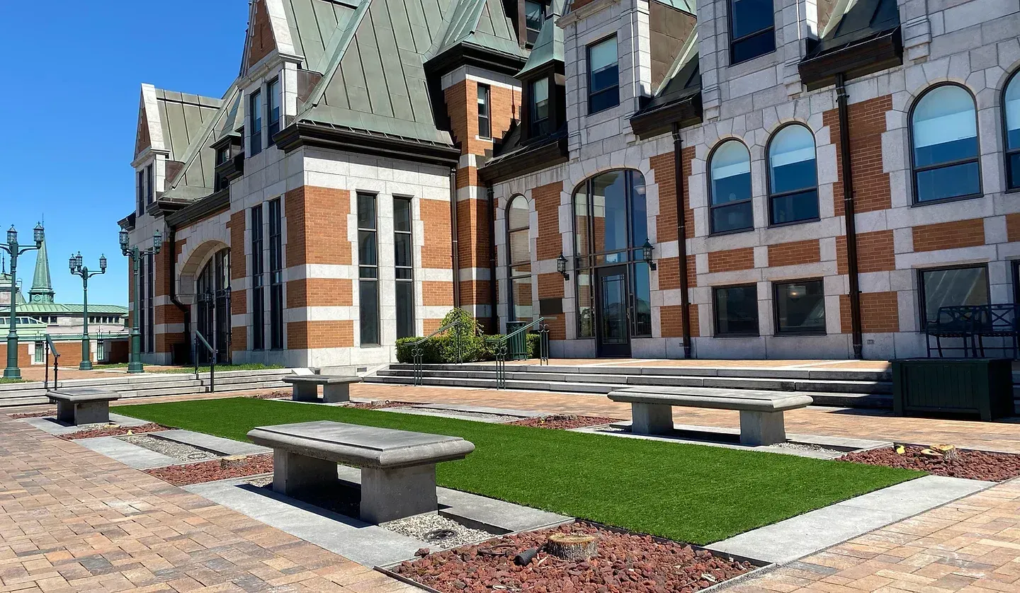 Une place extérieure lumineuse avec des allées de briques, des bancs en pierre et une pelouse rectangulaire devant un bâtiment historique.