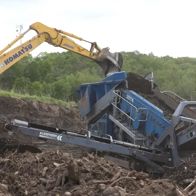 Une pelle mécanique Komatsu jaune charge de la terre et des débris dans une machine de criblage industrielle bleue à l'extérieur.