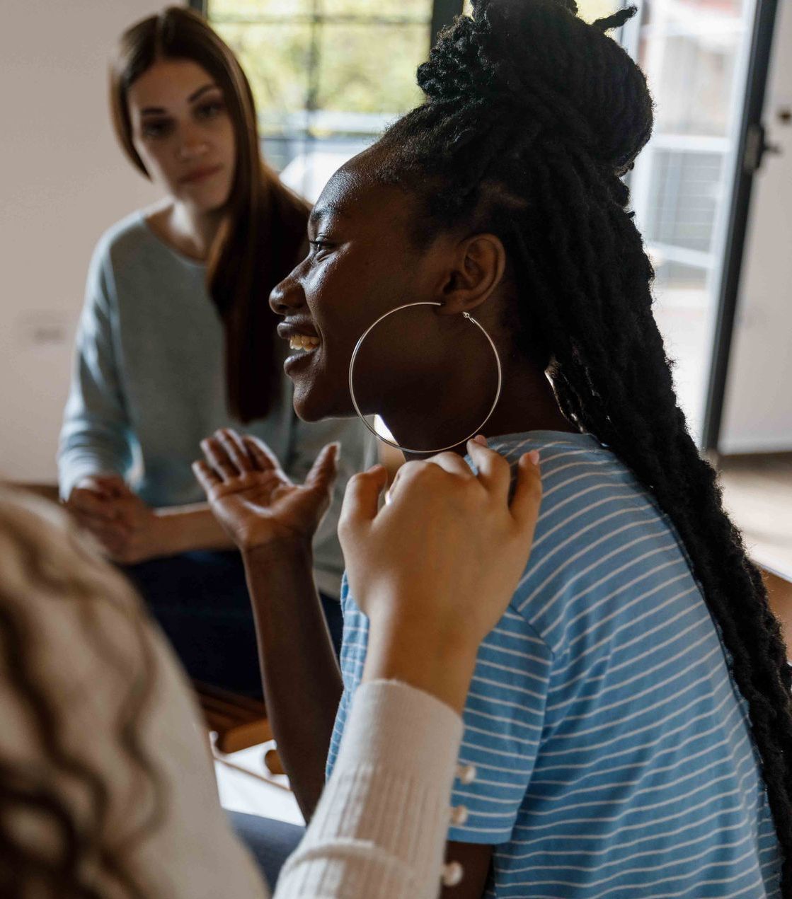 Black woman with braids speaks animatedly in a group, gesturing with hands. Others listen indoors.