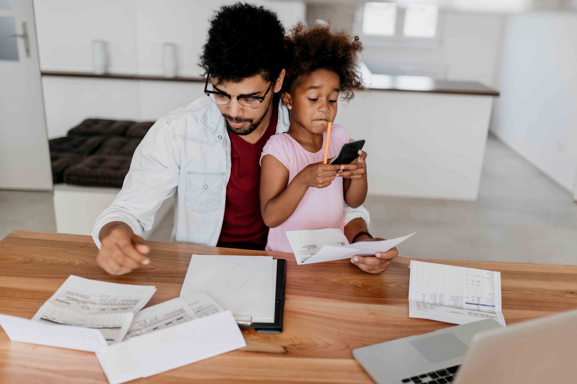 A man and a little girl are sitting at a table looking at papers and a cell phone.