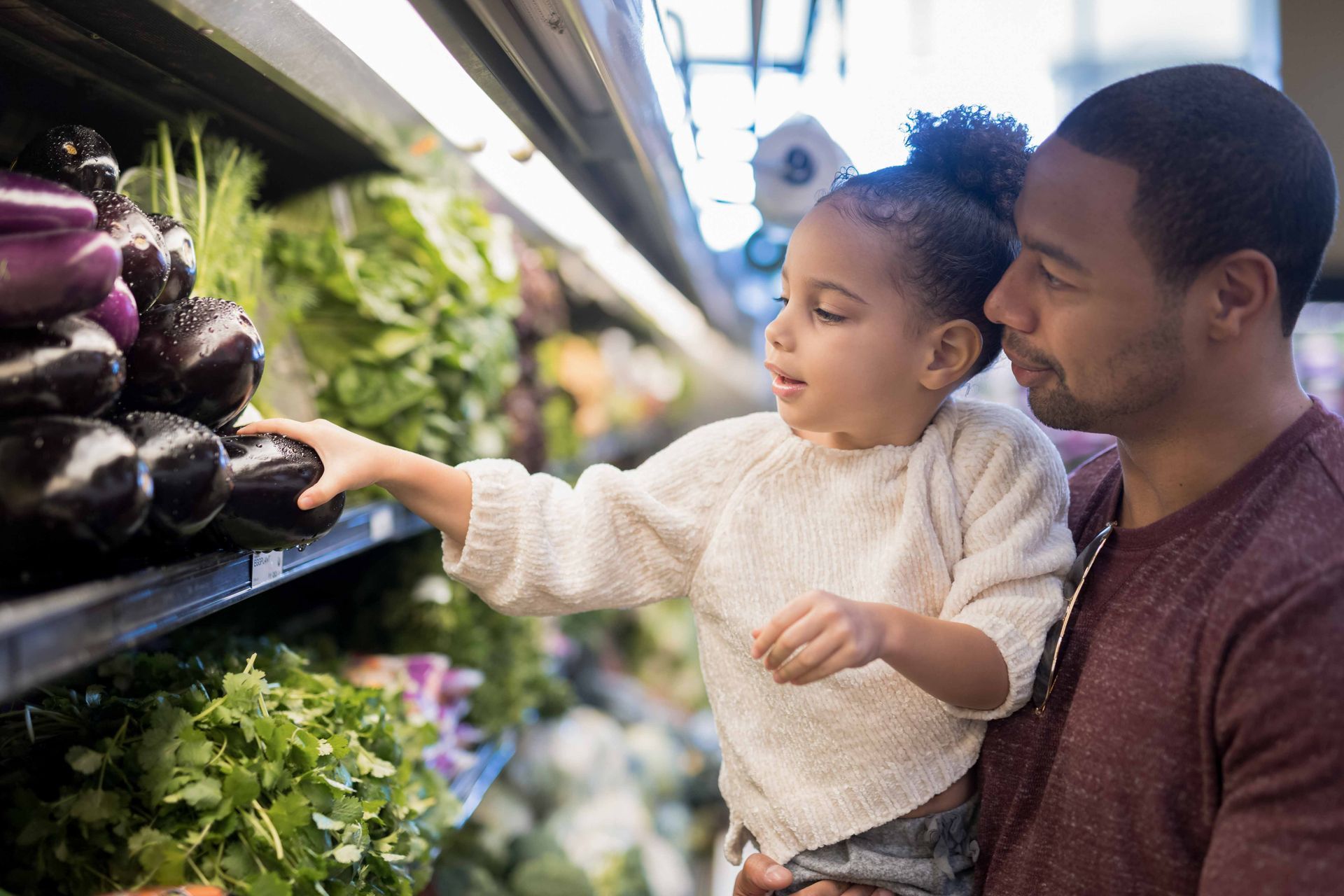 A father holds his young daughter as she reaches for an eggplant at a grocery store. They stand in front of a produce shelf.