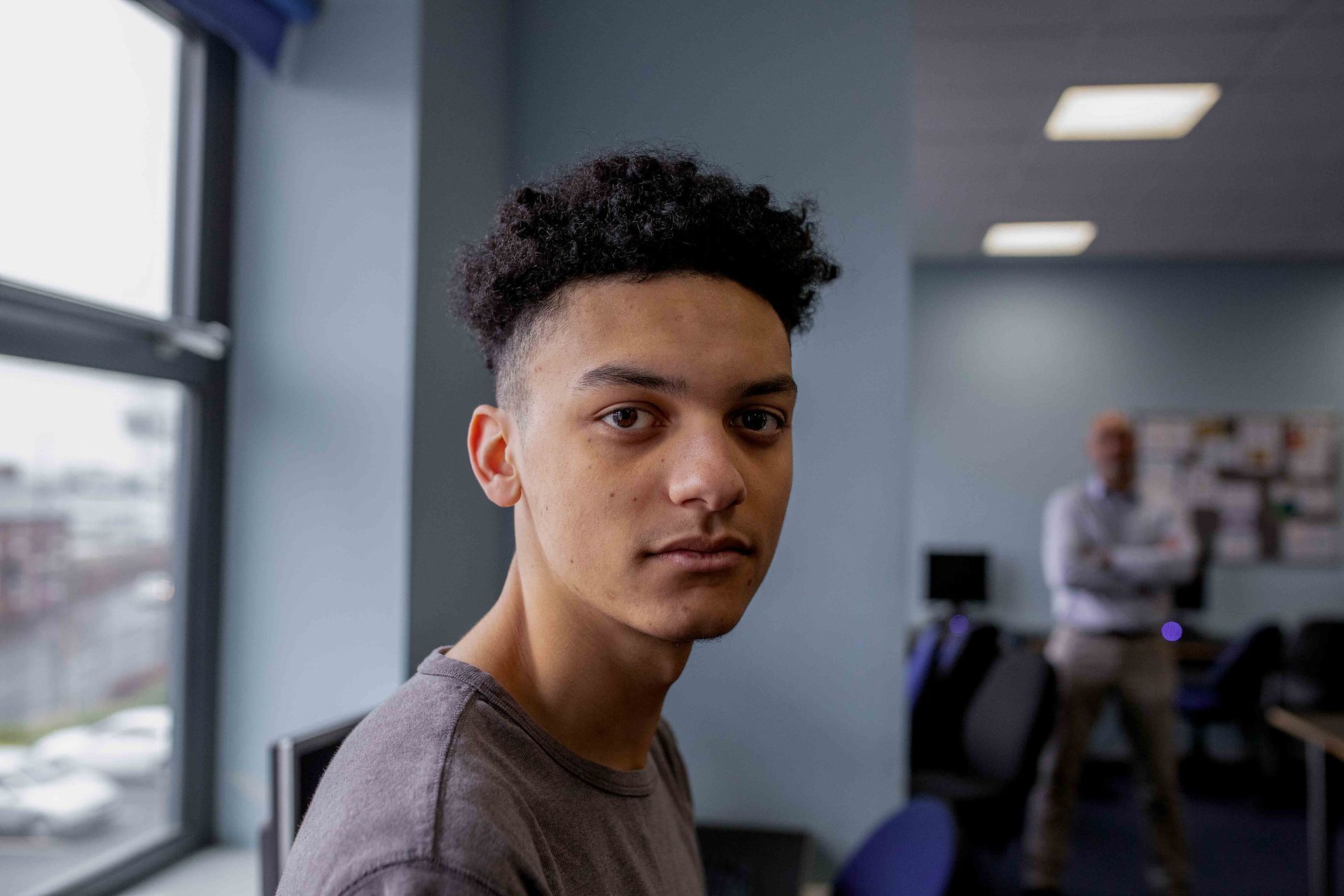 A young man is sitting in front of a window in a room and looking at the camera.