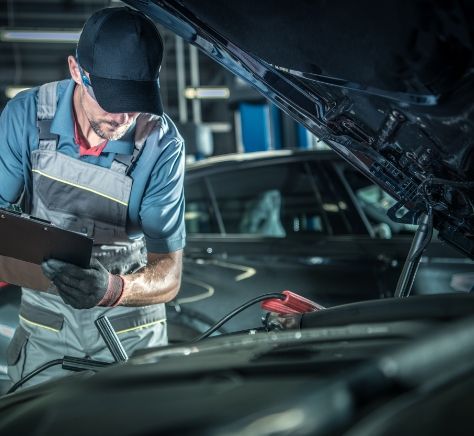 A man is working on a car with the hood open and looking at a clipboard.