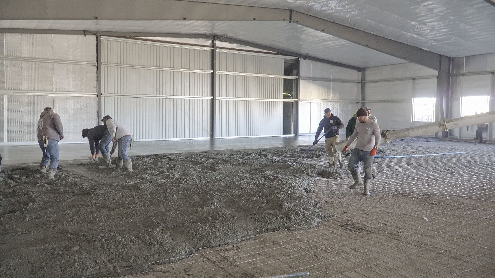 A person in orange gloves uses a long metal screed to level fresh concrete in a construction site setting.