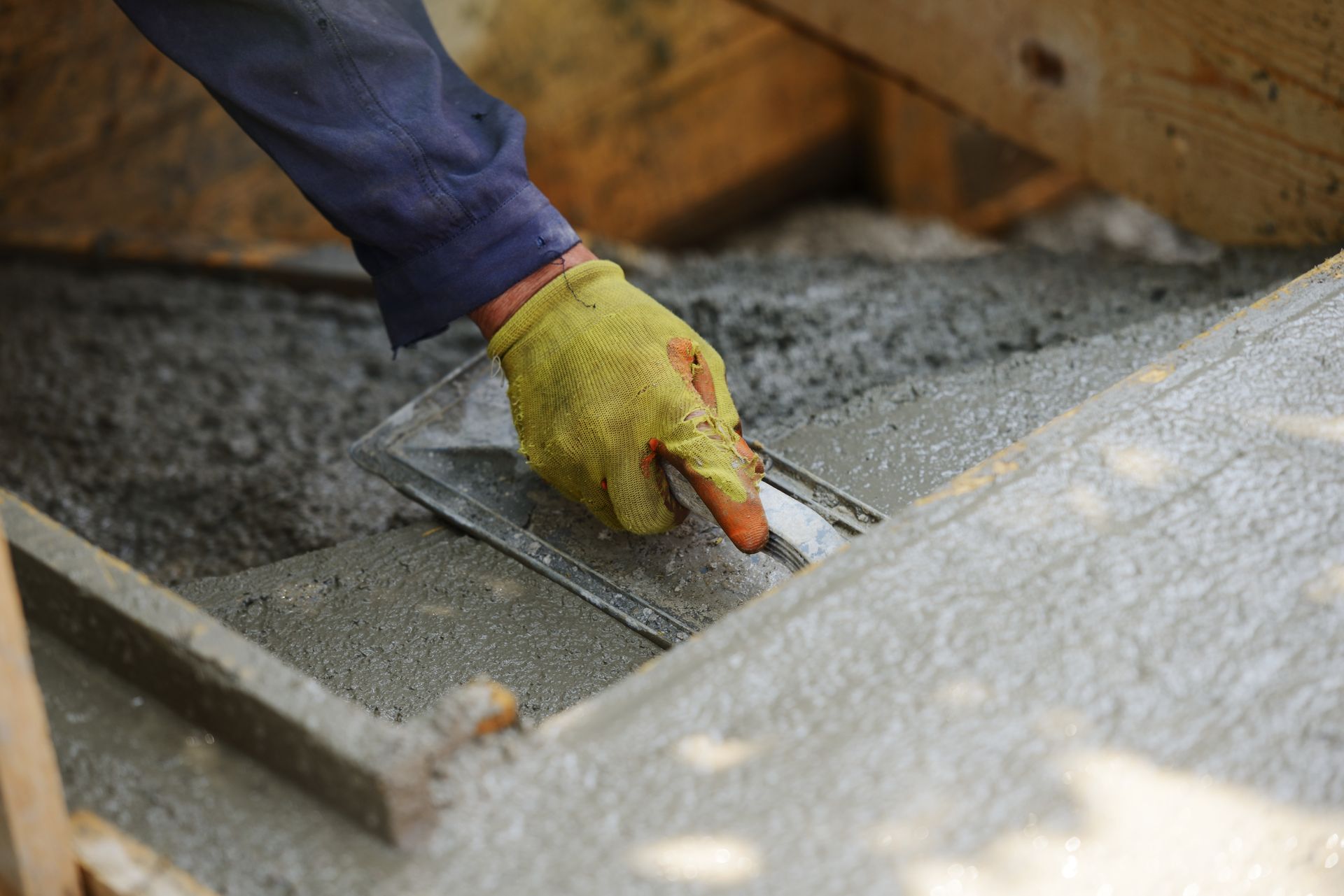 A gloved hand uses a metal trowel to smooth wet concrete inside a wooden formwork.