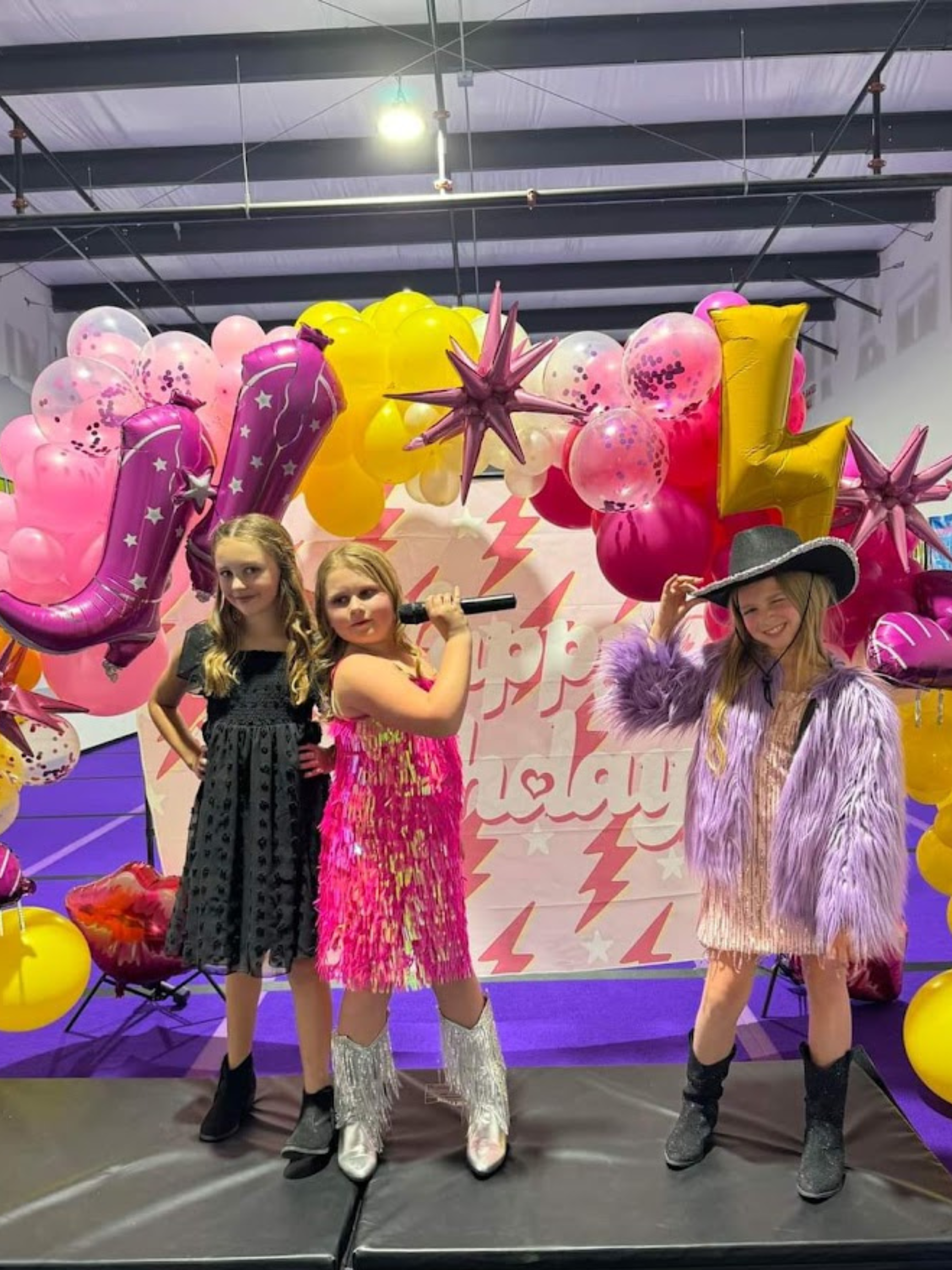 Three little girls are standing on a stage with balloons in the background.
