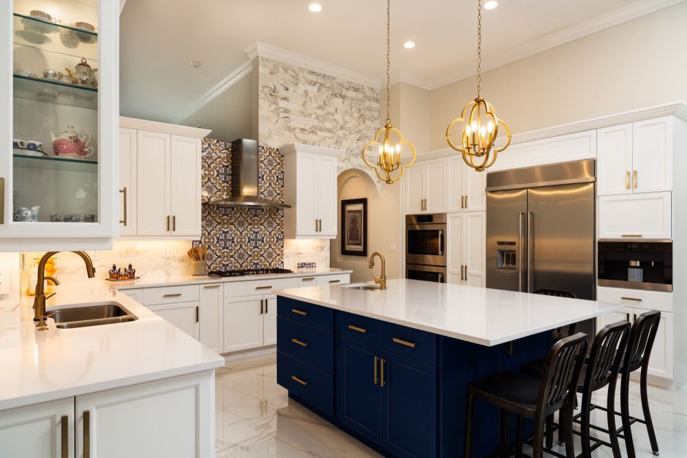 A kitchen with white cabinets, a large blue island, marble countertops, stainless steel appliances, and gold lighting.