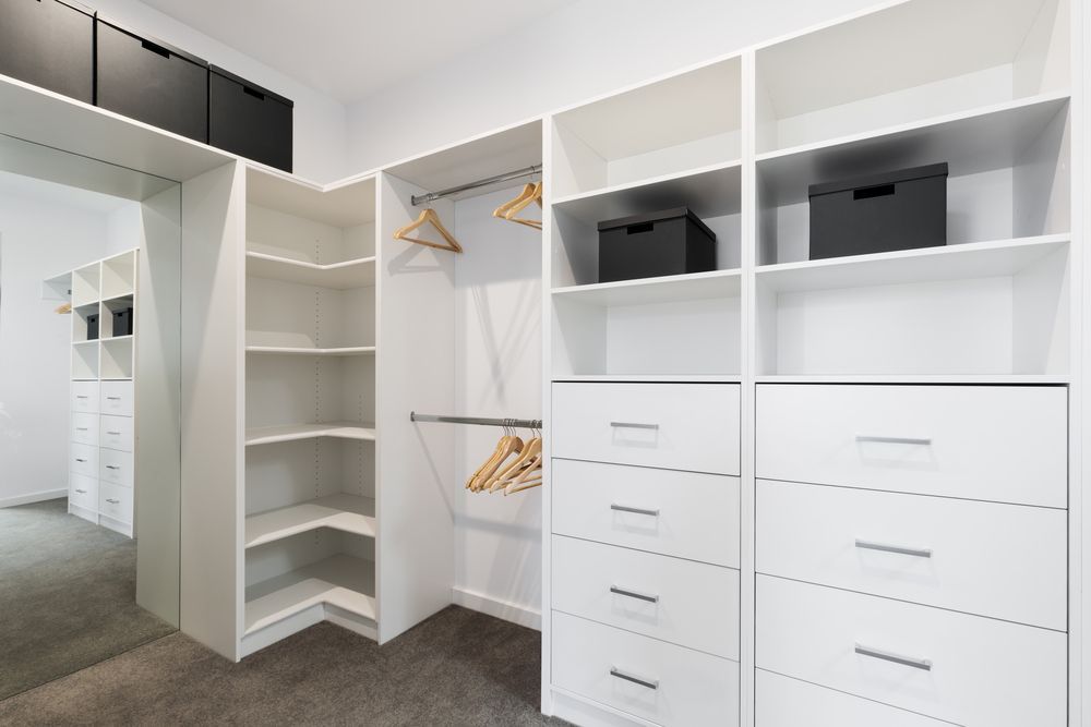 A clean, modern walk-in closet with white shelving, hanging rods, drawers, and black storage bins on a gray carpet.