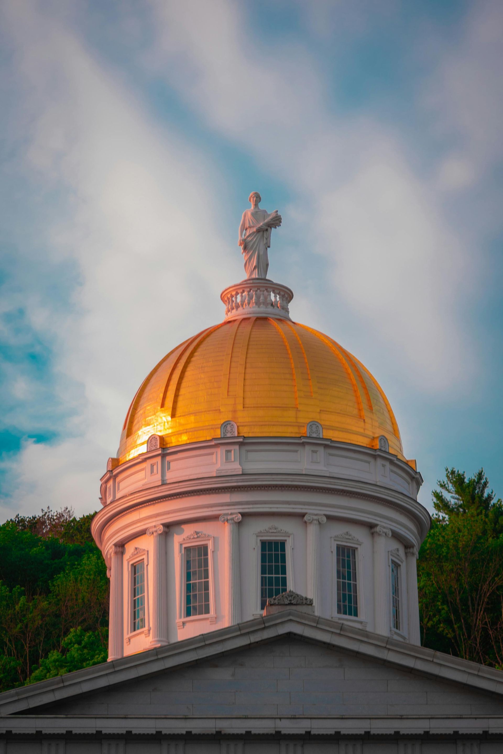 Golden dome of the Vermont State House with statue on top, set against a cloudy sky.