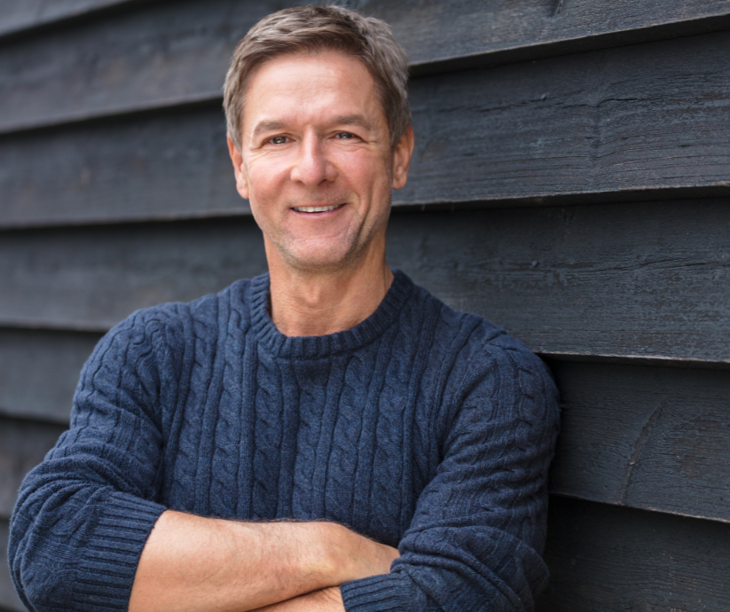 Man in blue sweater smiles, arms crossed, leaning against a dark wooden wall.