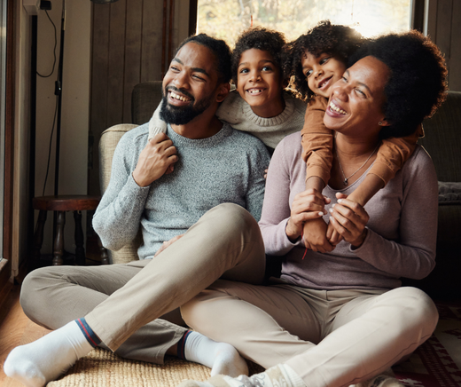 Family of four smiling, sitting on a floor. Two children hug their parents from behind; bright setting.