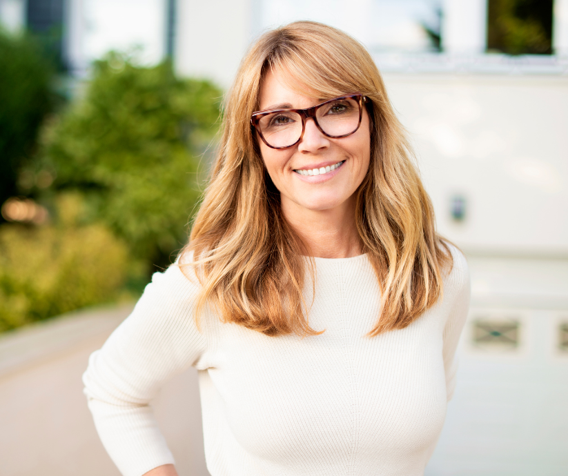 Woman with glasses smiles, standing outdoors in front of a building.