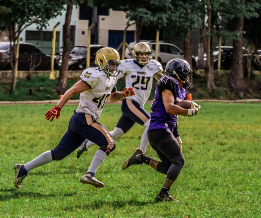 Football players in action on a grassy field, one running with the ball pursued by two opponents.