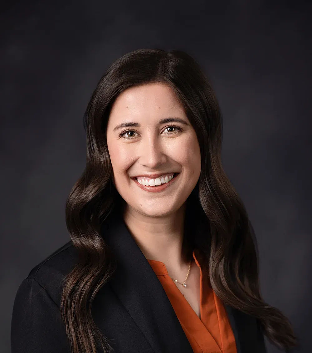 Woman with dark hair smiles, wearing a black blazer and orange top, against a dark gray background.
