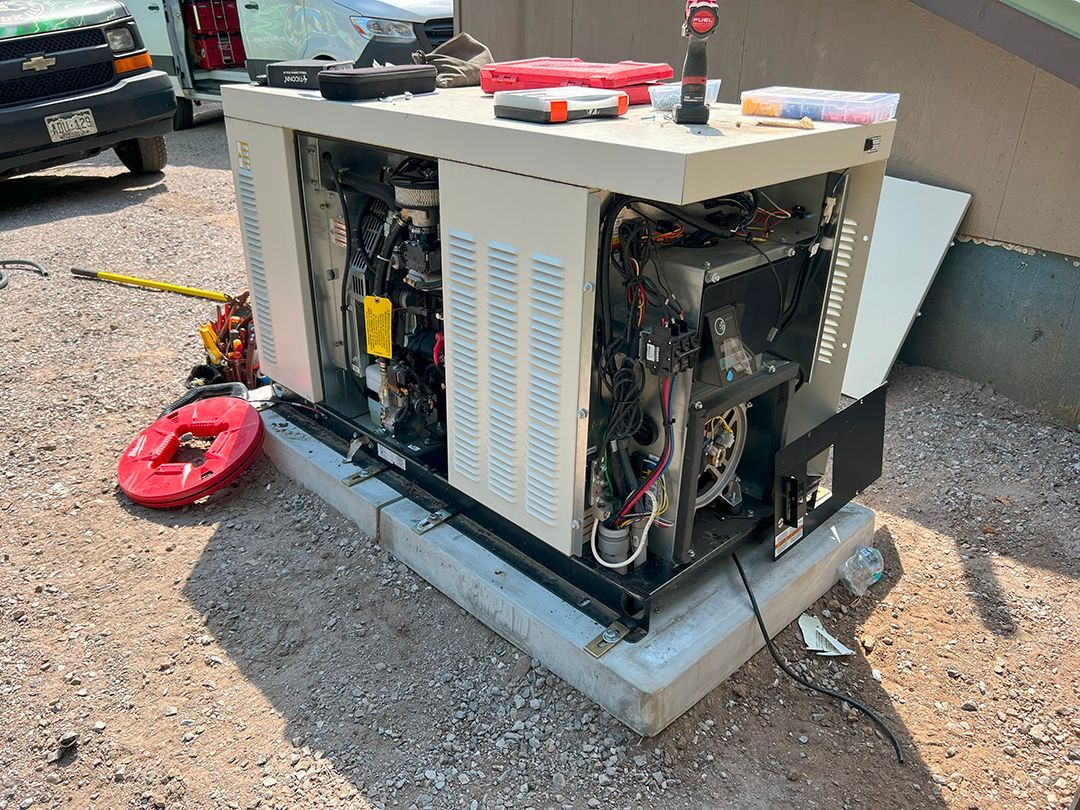 A beige home generator sits on a concrete base next to a house wall. A tool bag sits nearby.