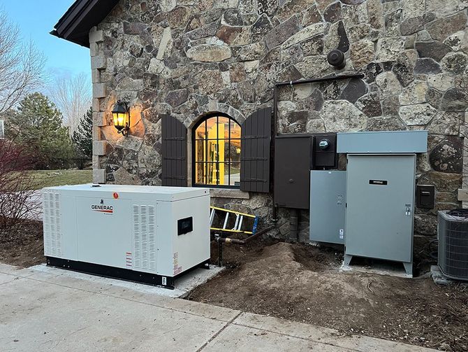 A beige Generac generator sits outside a building. Gray electrical box above it, brown leaves on the ground.