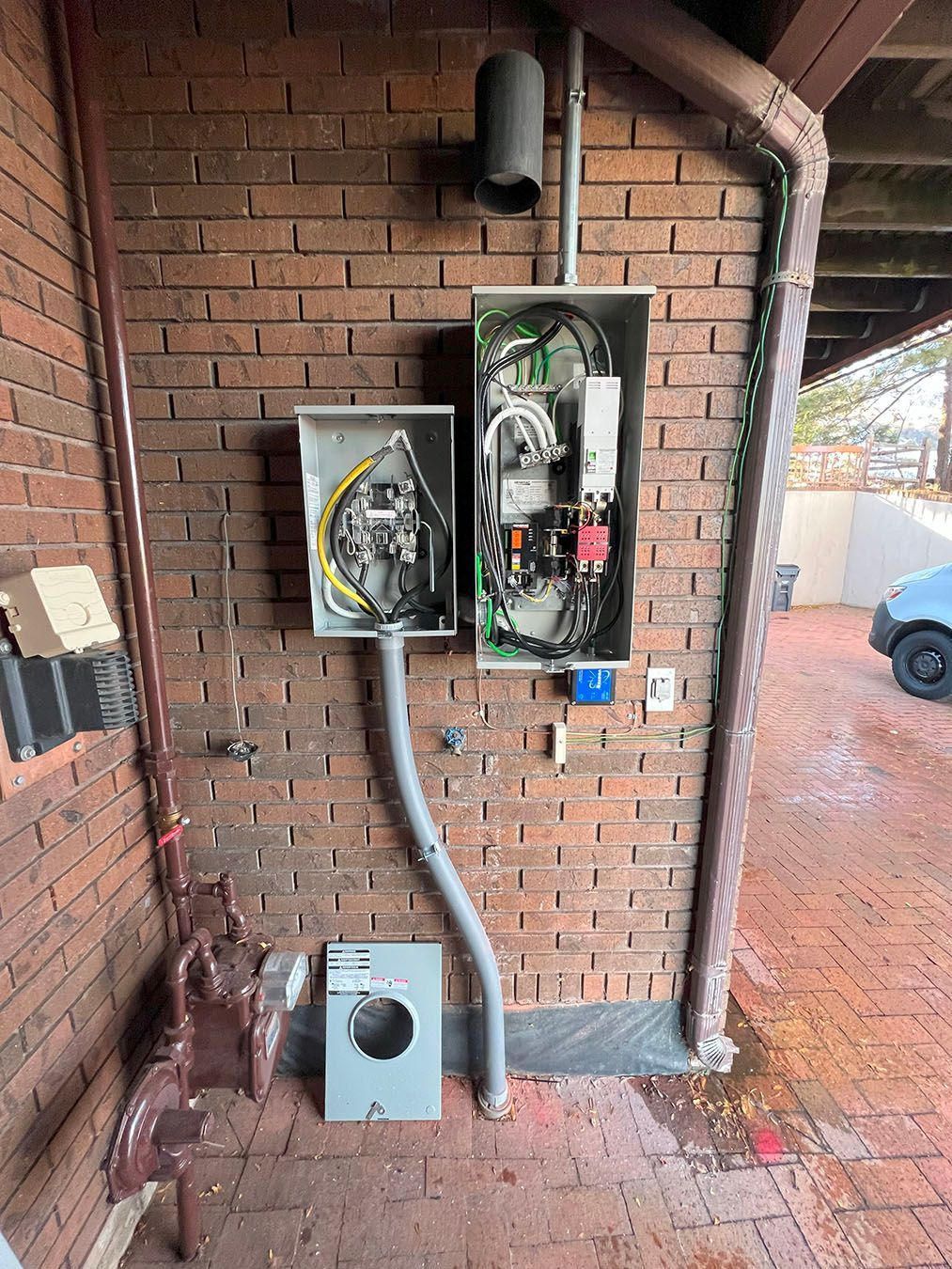 Electrical panel and meter box mounted on brick wall, with conduits and guttering visible.