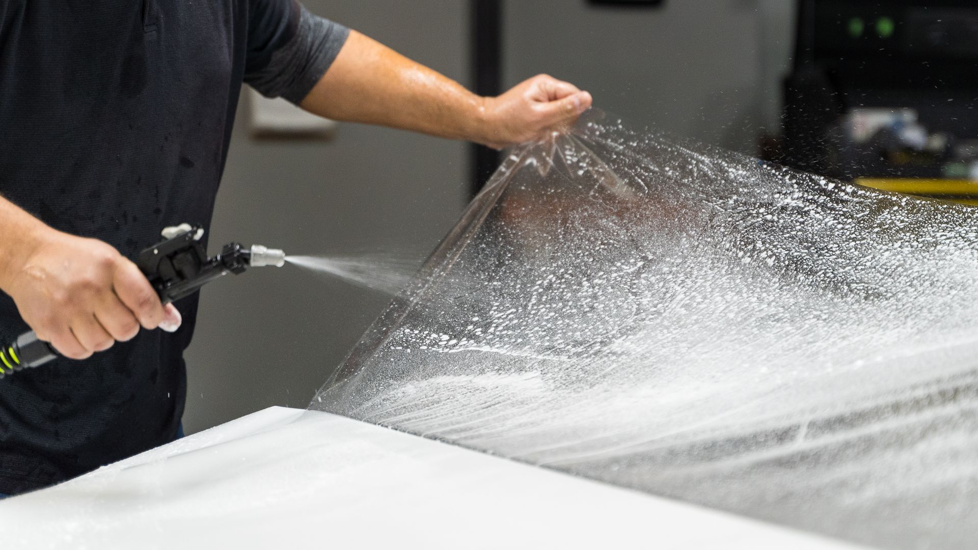 Person spraying water on a sheet of clear plastic on a white surface.