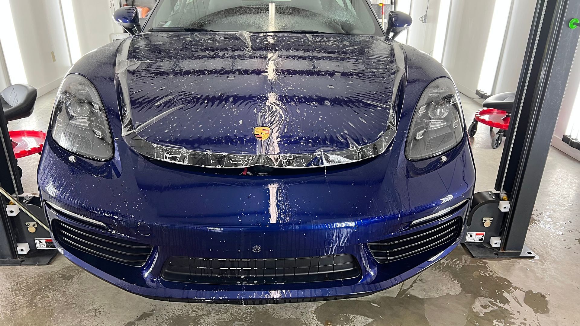 Blue Porsche being detailed, with soap on hood. Car is in a garage, on a lift.