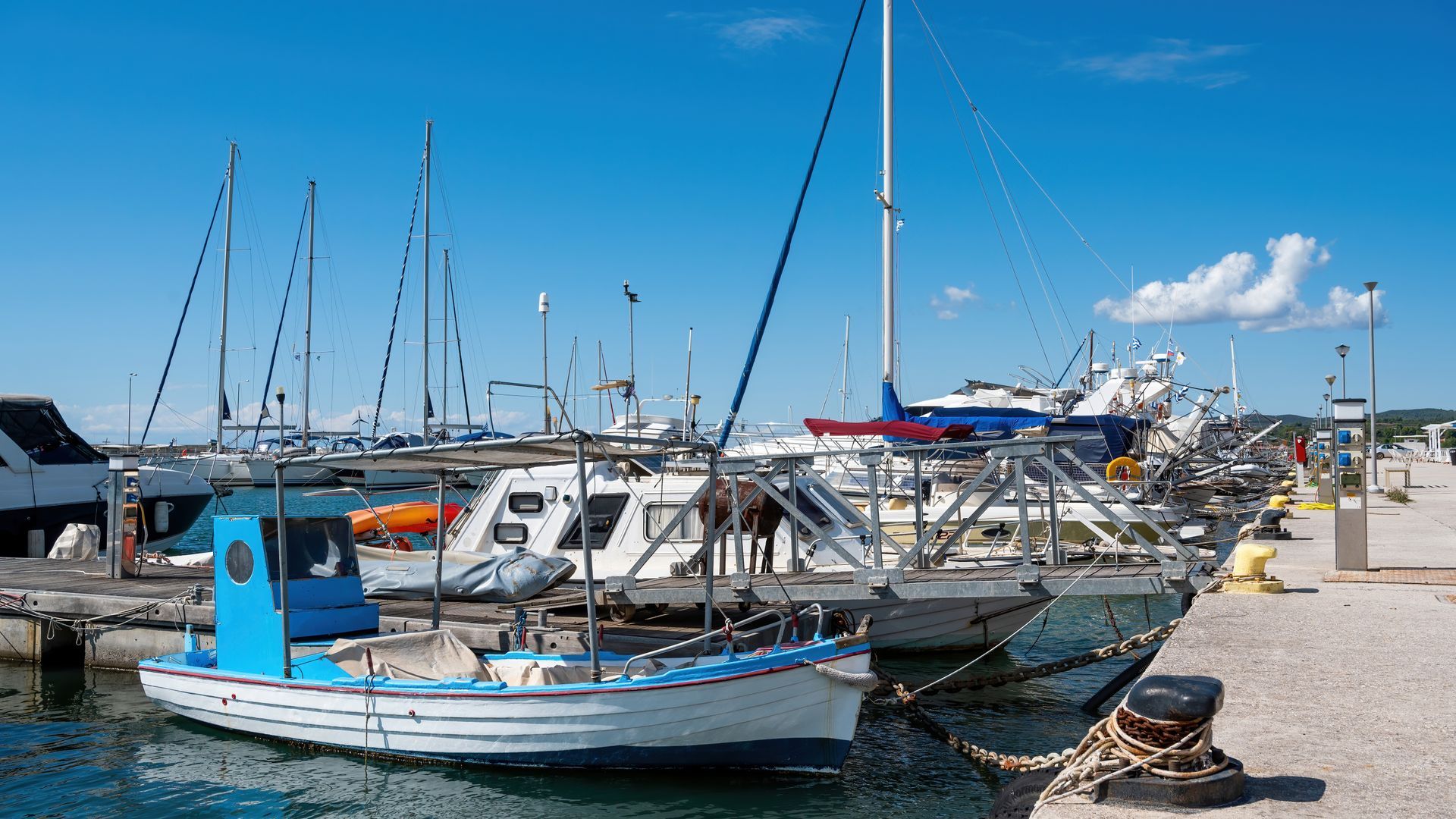 A small blue-and-white fishing boat moored at a harbor dock with several sailboats and motorboats docked nearby.