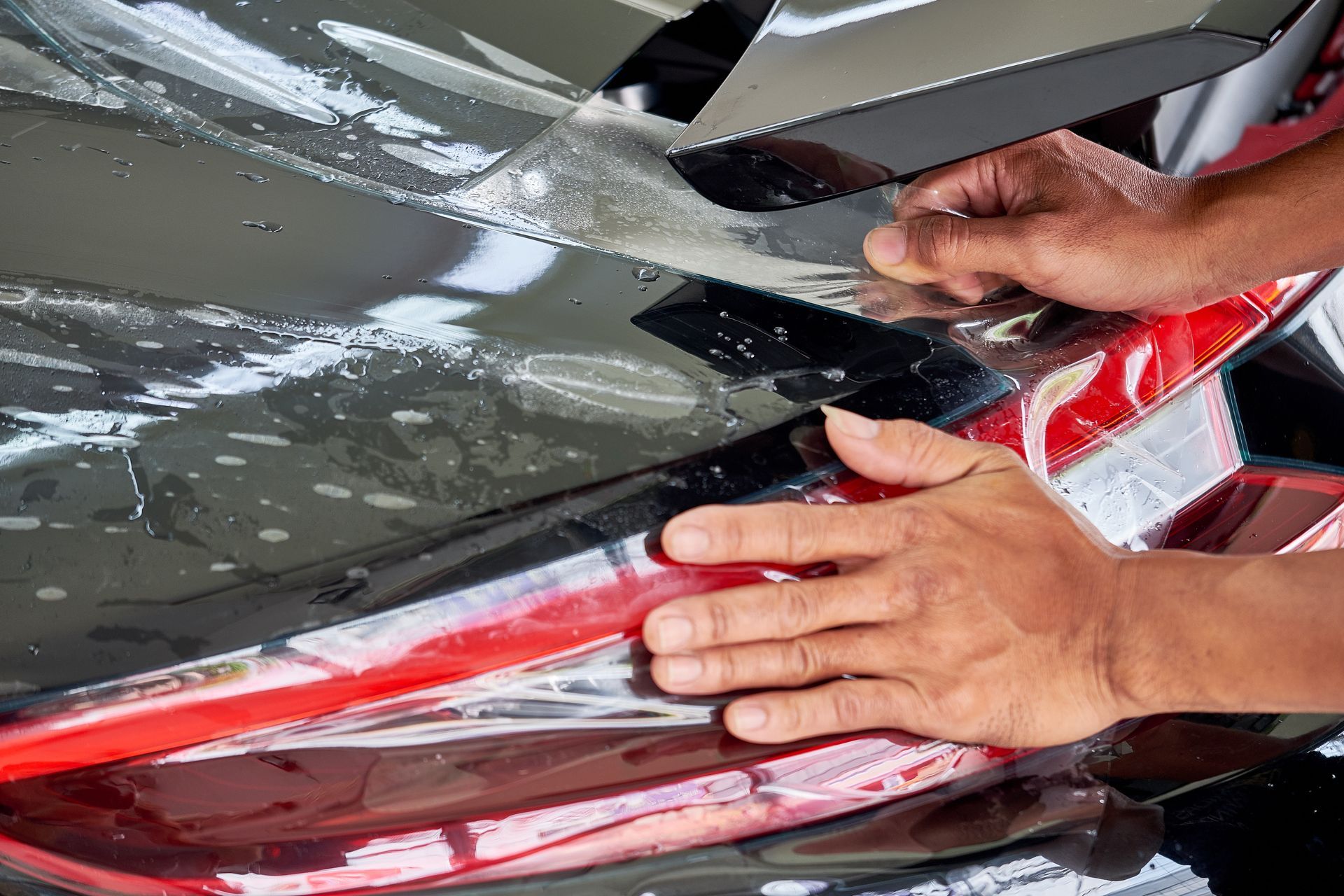Hands applying protective film to a car's taillight, using a tool to smooth the surface.