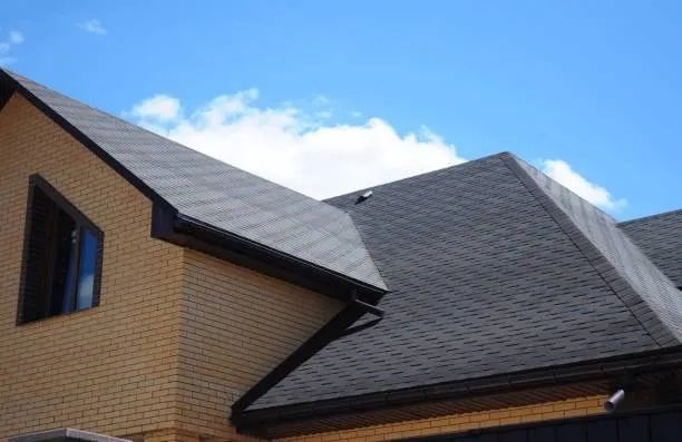 Beige brick house with dark gray shingle roof and black gutters against a blue sky.