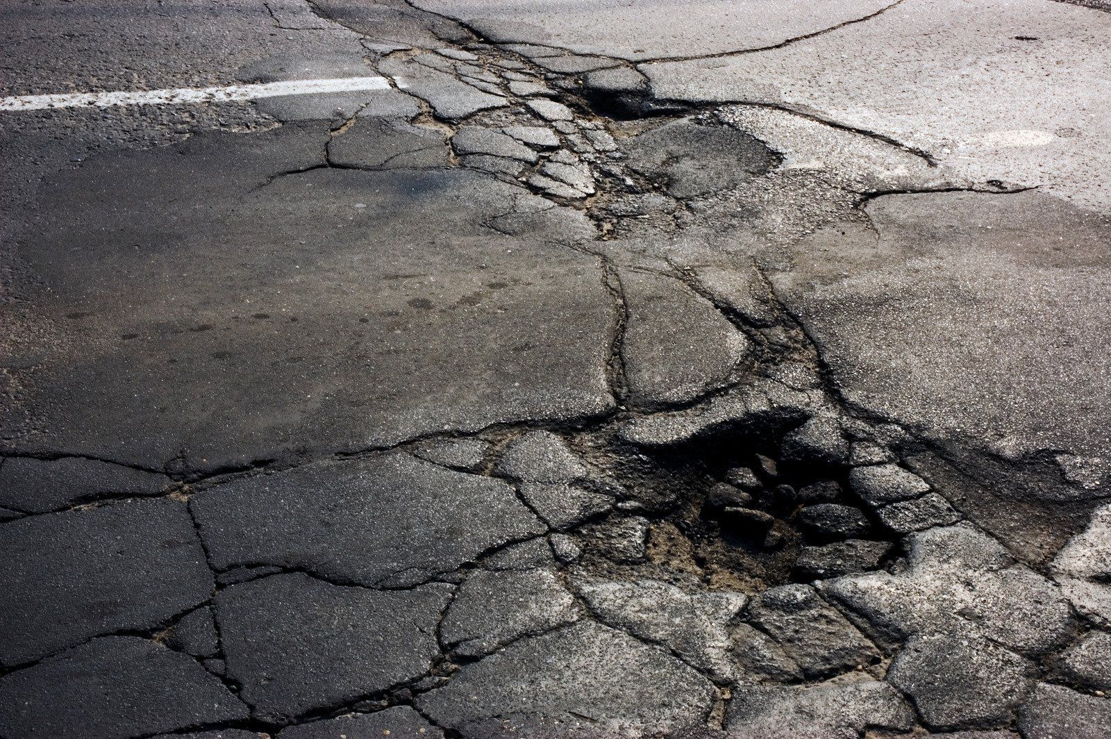 Cracked and damaged asphalt road surface, showing a pothole and large cracks.