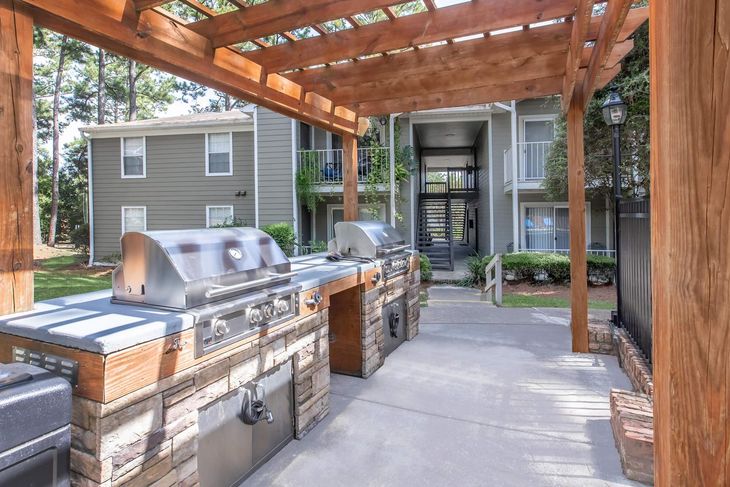 Outdoor grilling station with grills under a wooden pergola, apartment buildings in background.