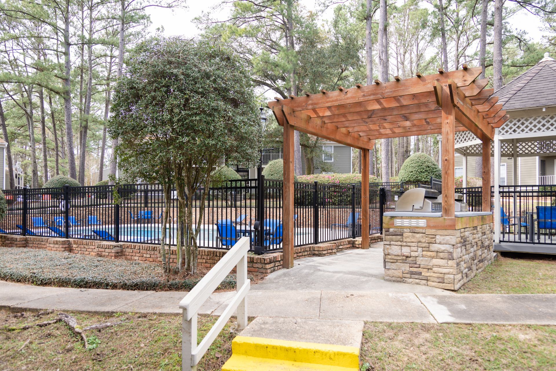 Pool area with pergola, grill, and wrought-iron fence. Blue pool chairs are visible.
