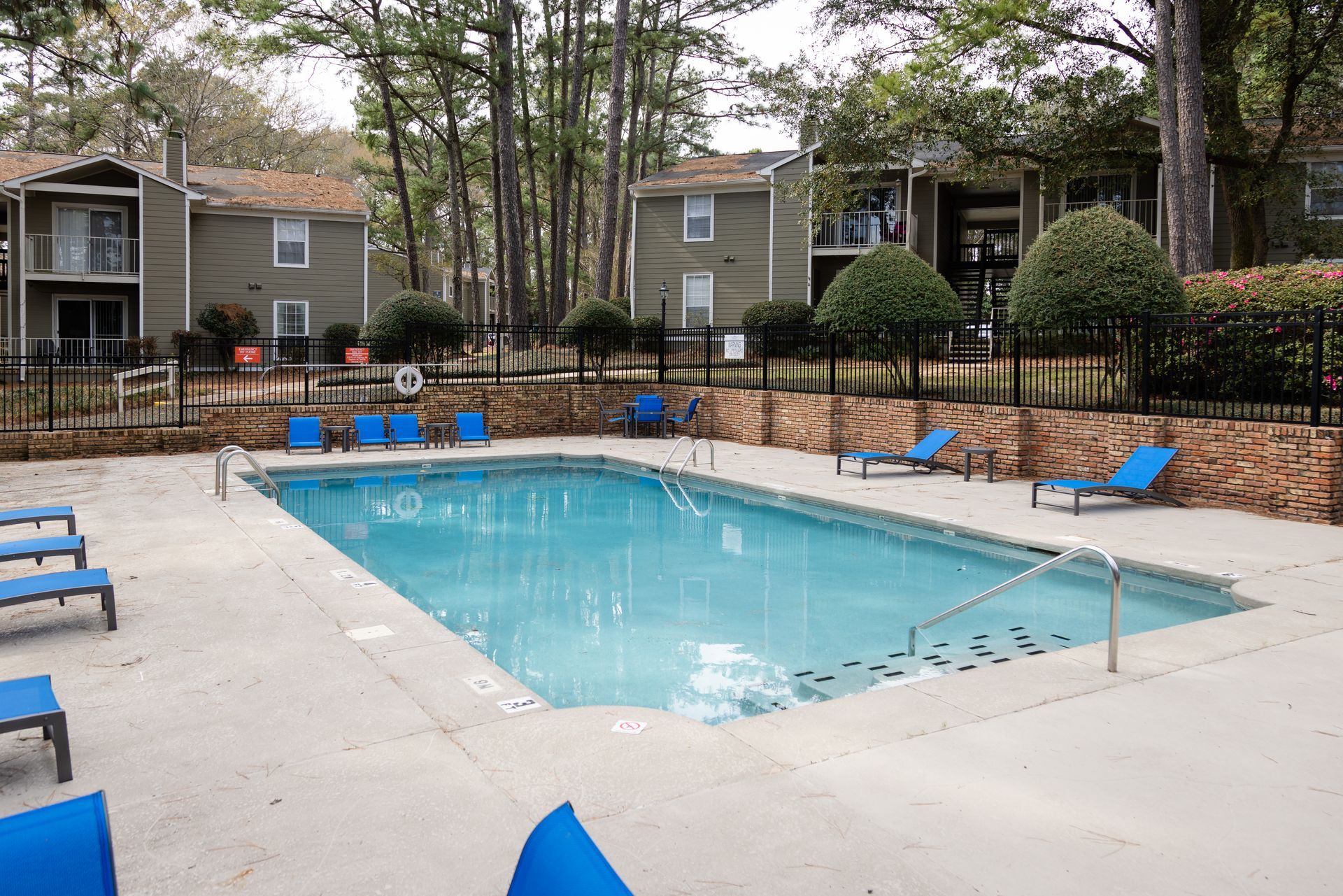 Swimming pool with blue lounge chairs, near two-story apartment buildings, in a wooded area.
