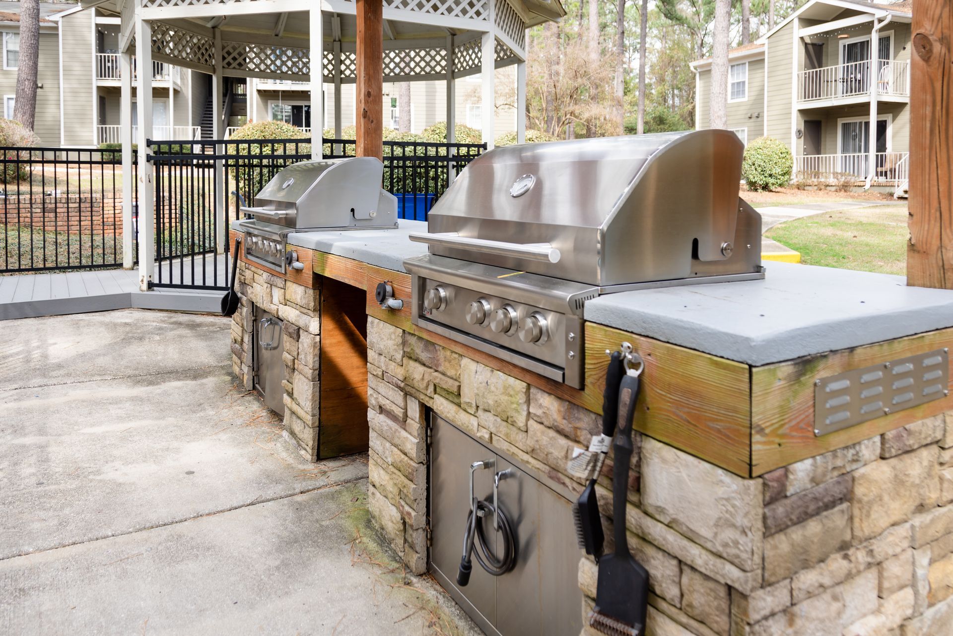 Outdoor grilling station with stainless steel grills, stone facade, and a gazebo in the background.