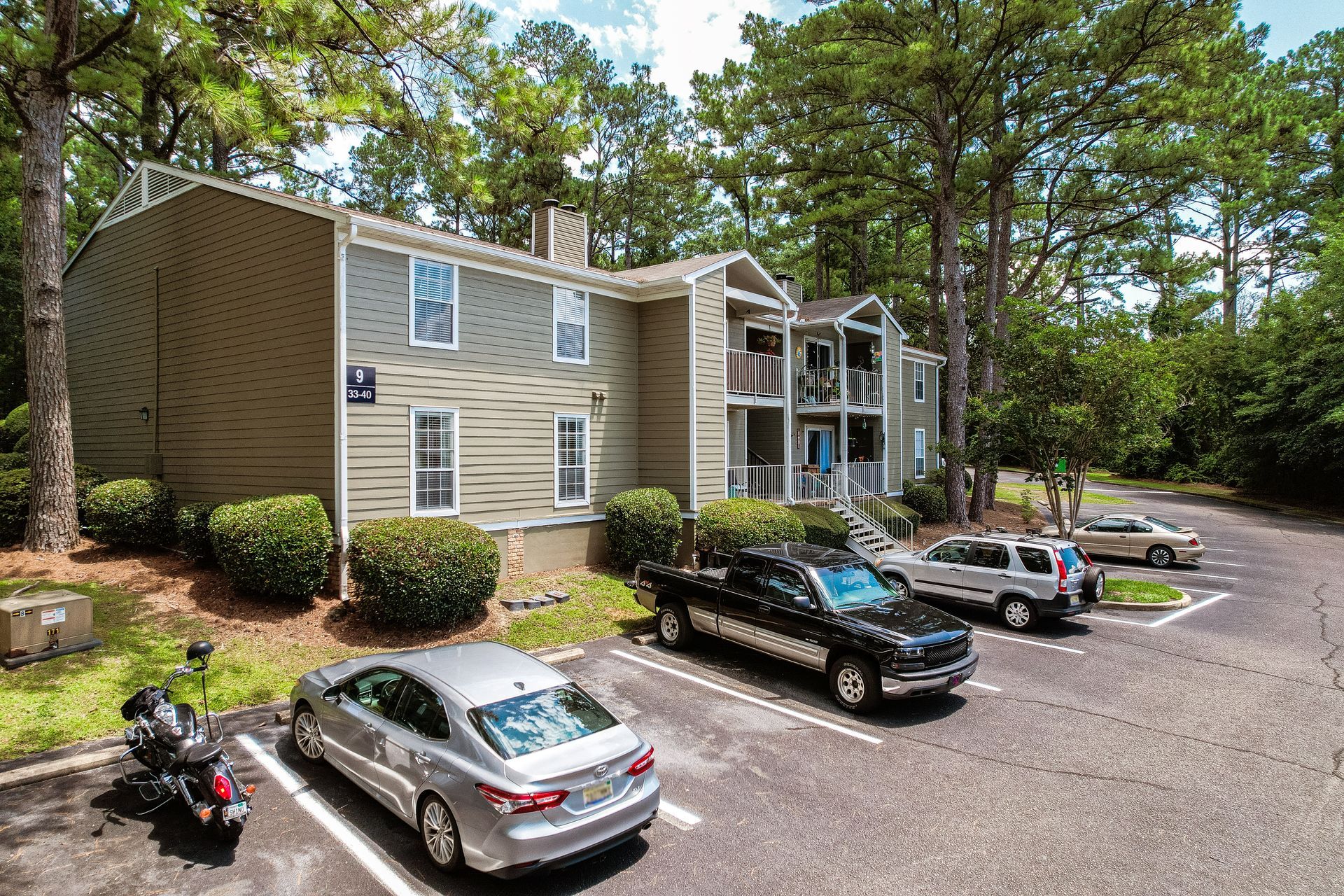 Apartment building exterior with parked cars. Light brown siding with green trees.