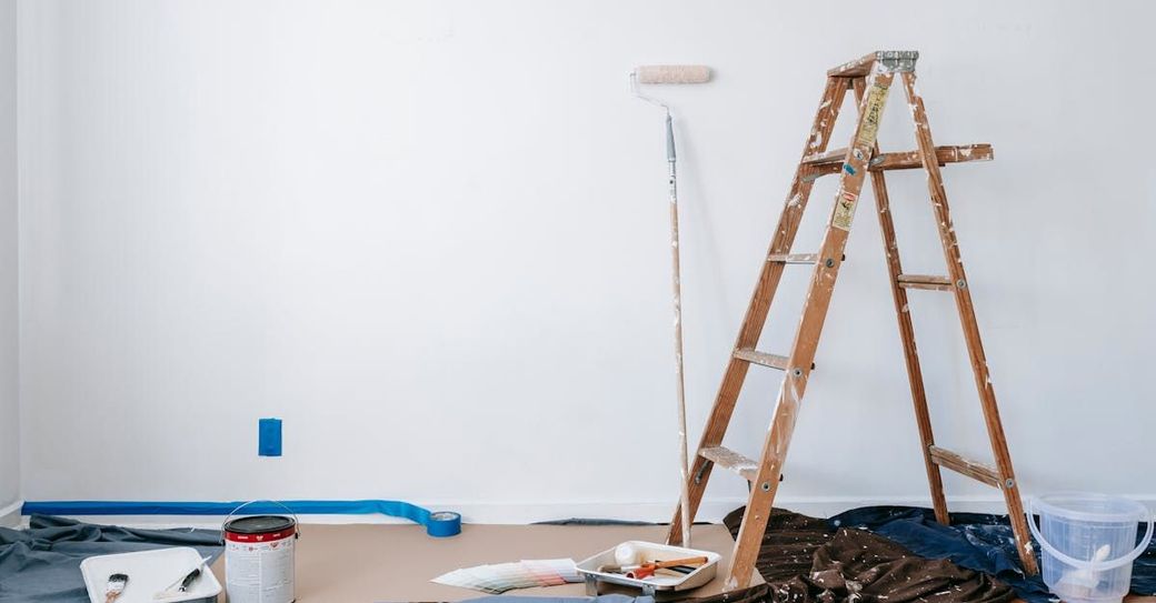 Ladder, paint roller, and supplies in a room with white walls, indicating a painting project.