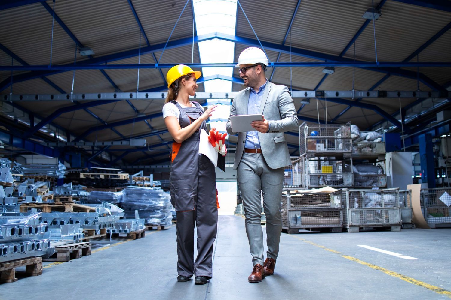 Woman in yellow hard hat and overalls with man in a suit, both walking and talking in a factory setting.
