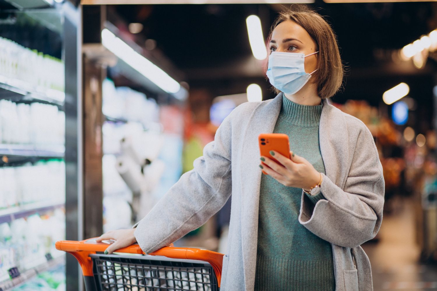 Woman in a face mask uses a phone while shopping in a grocery store, holding a shopping cart.
