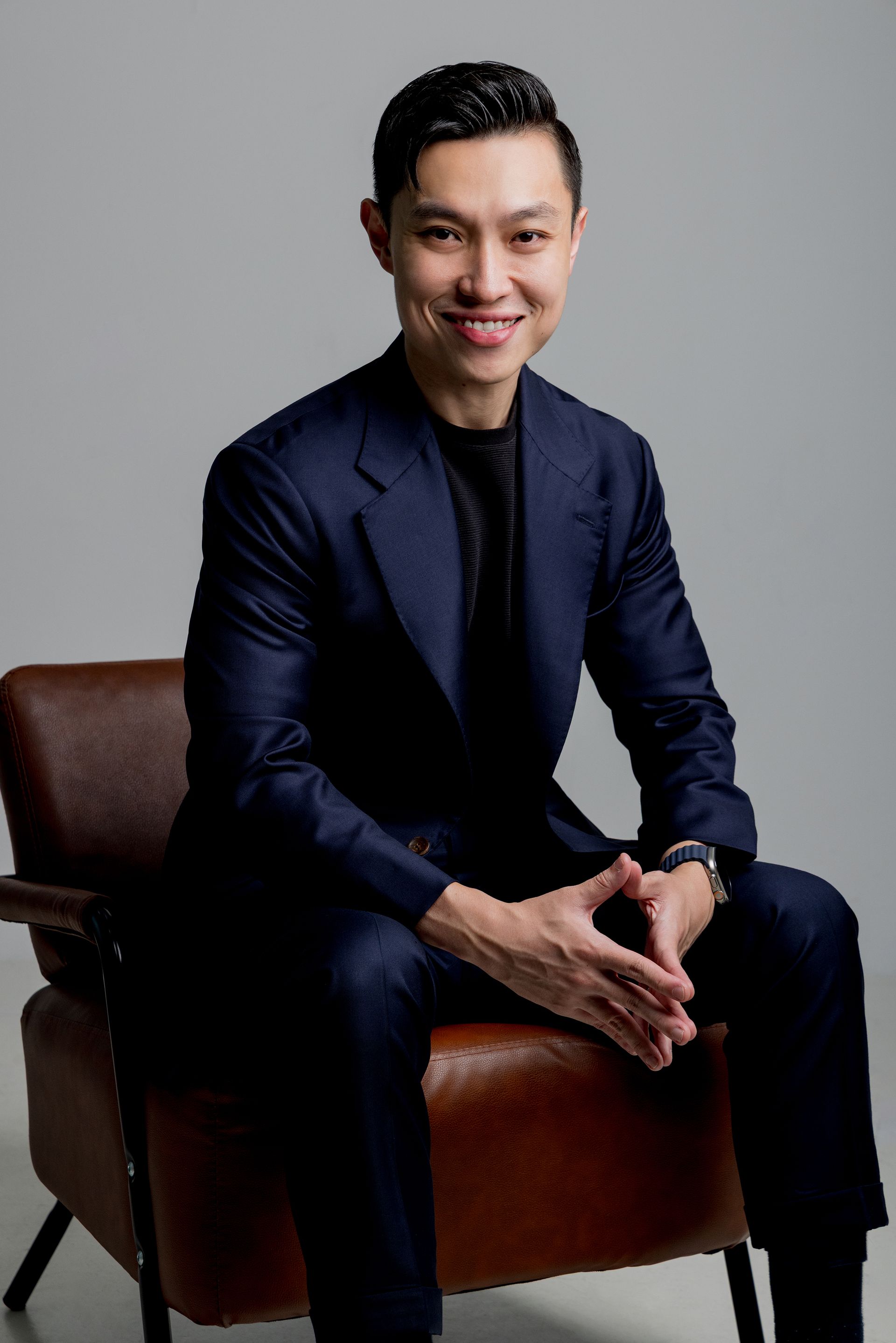 Man in a navy suit sits in a brown armchair, smiling with hands clasped. Light background.