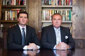 Two men in suits and ties are sitting at a table in front of a bookshelf.