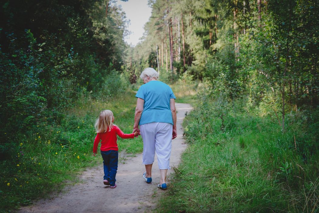 Two people walking hand in hand on a wooded trail, surrounded by green trees and grass.