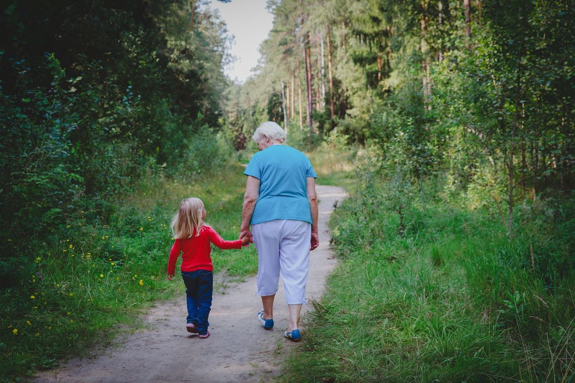Two people walking hand in hand on a wooded trail, surrounded by green trees and grass.