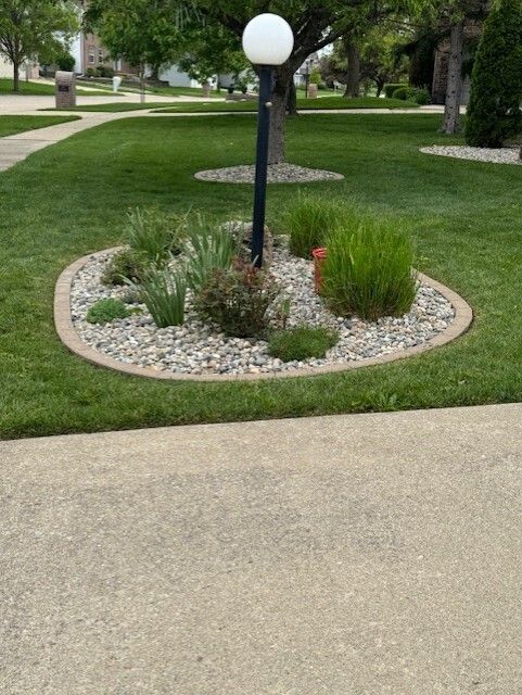 Flower bed around a lamp post with greenery, rocks, and a tan border on a green lawn.