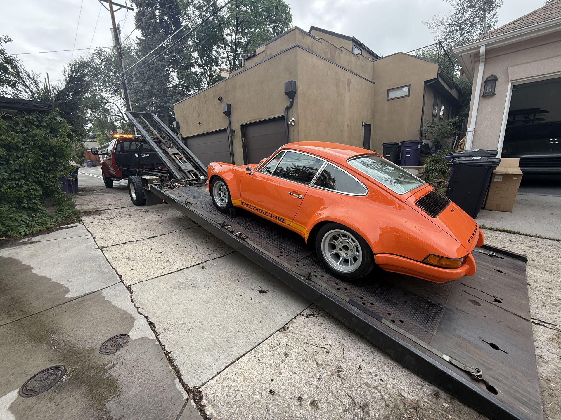Orange Porsche 911 being loaded onto a flatbed tow truck in front of a house with a garage.