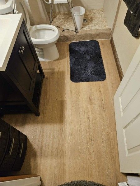 Bathroom with wood-look flooring, dark vanity, toilet, and black bath mat.