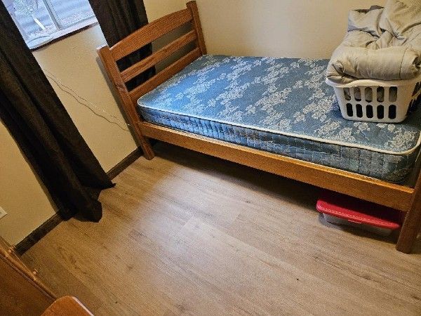 Wooden bed frame with blue floral mattress, laundry basket on the bed, brown curtains, and light wood floors.