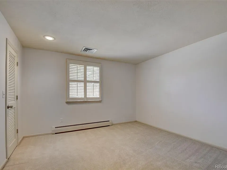 Empty, white-walled room with closed door, window with shutters, and beige carpet.