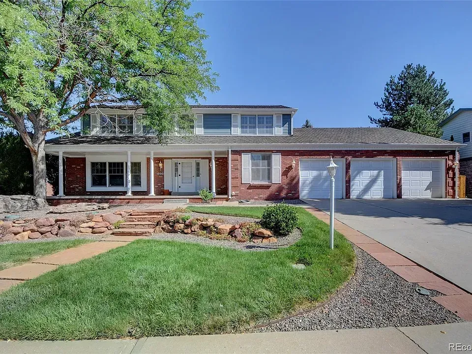 Two-story brick house with covered porch, three-car garage, and landscaped yard on a sunny day.
