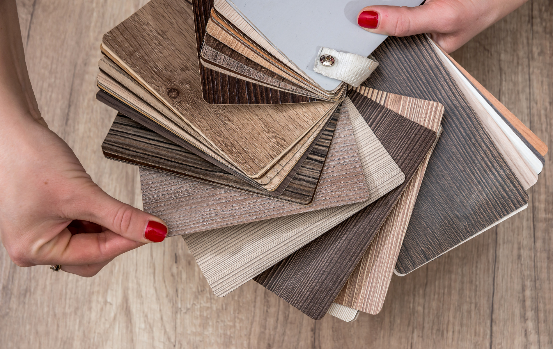 Hands holding a fan of wood flooring samples in various neutral tones, on a wood surface.
