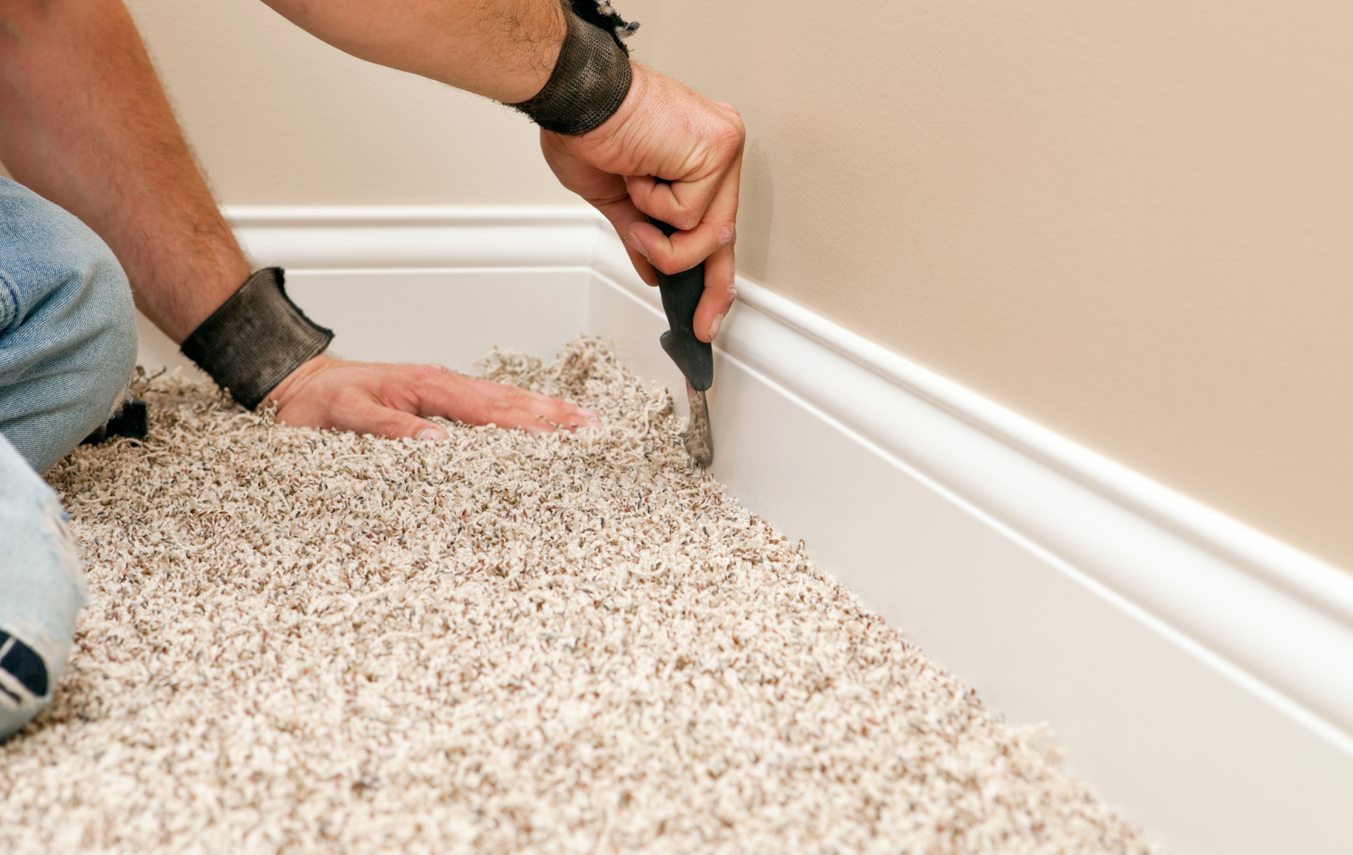Person using a tool to trim carpet along a white baseboard, near a beige wall.